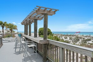Eine Holzterrasse mit Stühlen und einer Pergola mit Blick auf einen Strand mit Sonnenschirmen.