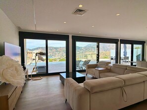 Ein modernes Wohnzimmer mit einer Couch, einem Couchtisch und einem Fernseher. Große Fenster bieten Blick auf einen Hügel und einen Pool.