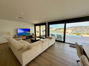 Ein modernes Wohnzimmer mit einem großen Flachbildfernseher, einem beigefarbenen Ecksofa und einem Essbereich mit Blick auf einen Swimmingpool und die Skyline.