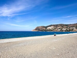 A beach with a pebble shore, a clear blue sky, and a distant hill with buildings. - Casa Montemar (Almuñécar)
