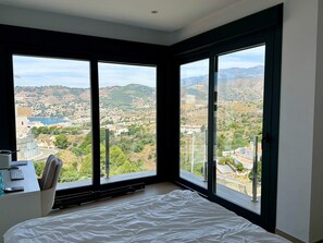 Ein Schlafzimmer mit einem großen Bett, einem Schreibtisch mit Stuhl und Blick auf eine hügelige Landschaft durch große Fenster.