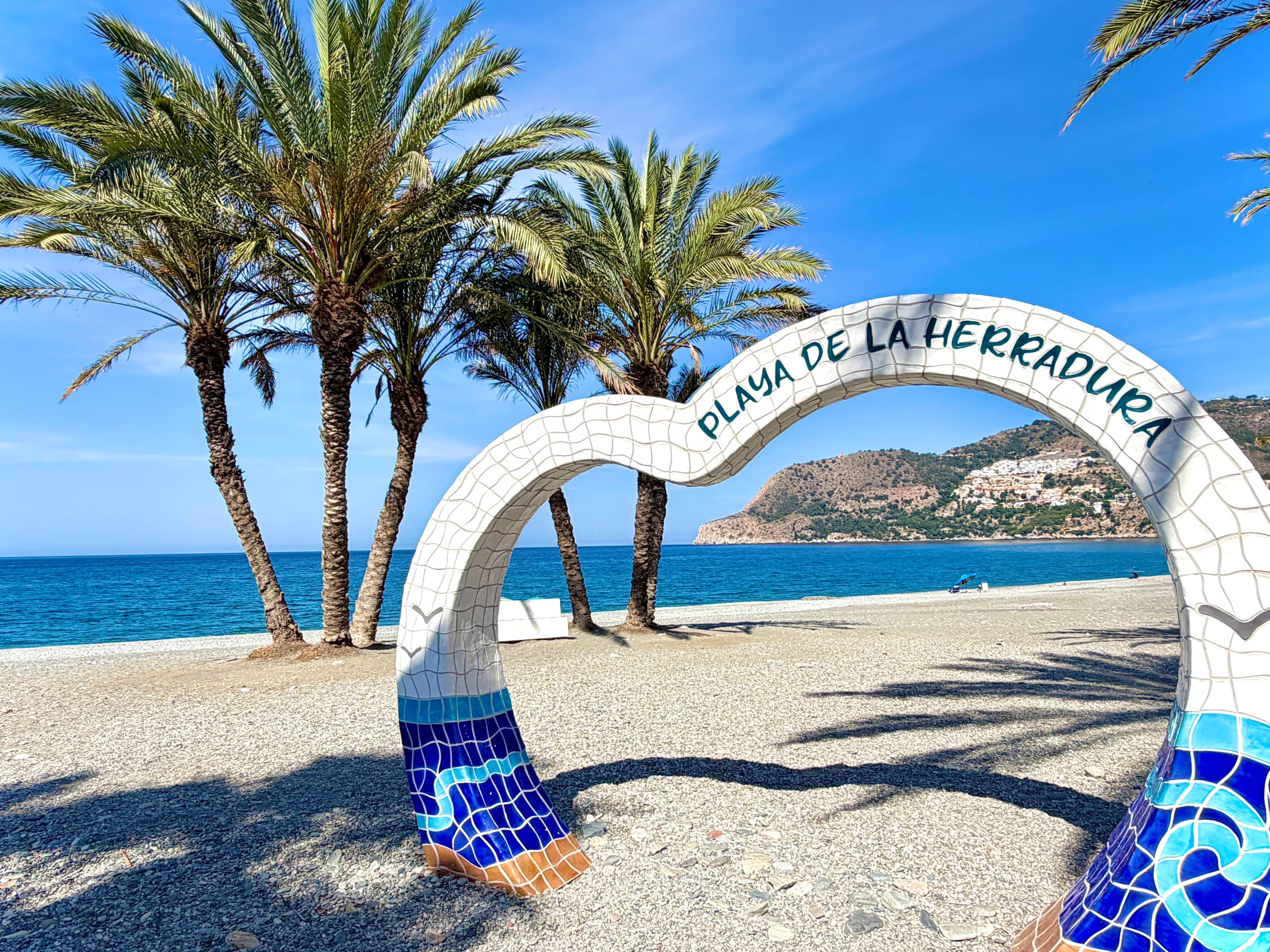 A beach entrance arch with 'PLAYA DE LA HERRADURA' text, palm trees, and a clear blue sky.