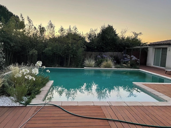 Ein rechteckiger Pool mit Holzterrasse im Vordergrund, umgeben von dichtem Grün und einem Haus im Hintergrund.