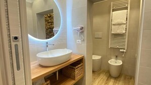 A bathroom with a round mirror, white sink, wooden vanity, wicker baskets, towel rack, and a bidet.
