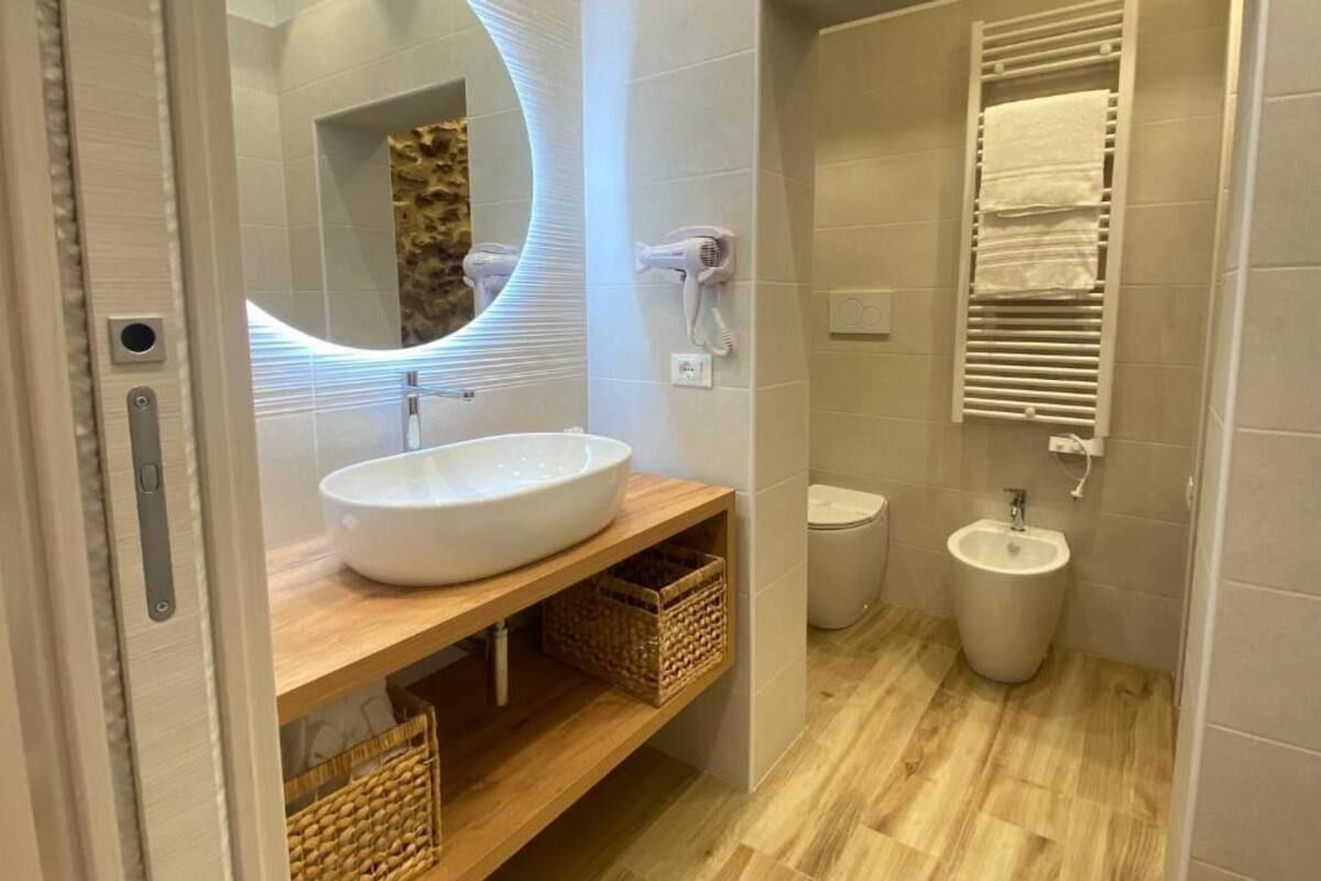 A bathroom with a round mirror, white sink, wooden vanity, wicker baskets, towel rack, and a bidet.