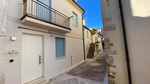 A narrow street with a building featuring a white door, a balcony with a metal railing, and a stone wall.