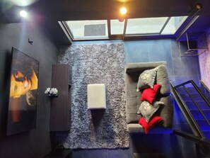 Una habitación con un sofá gris, un pequeño reposapiés blanco, un televisor montado en la pared y un tragaluz.
