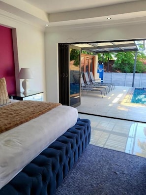 Un dormitorio con una cama grande, una mesita de noche con lámpara y vistas a una zona de piscina con sillones de relax.
