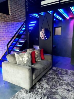 A modern living room with a grey sofa, red and silver cushions, a round mirror, and a staircase with blue lighting.