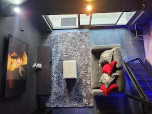 A room with a grey couch, a small white ottoman, a wall-mounted TV, and a skylight.