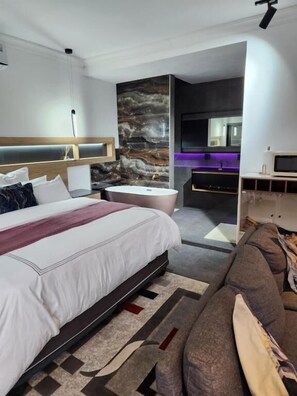A modern hotel room with a large bed, a bathroom with a bathtub, a sofa, and a TV.