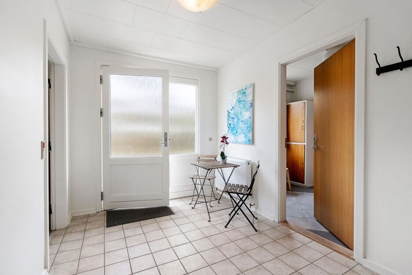 A hallway with a tiled floor, a door with frosted glass panels, a small table with a chair, and a wooden door on the right.