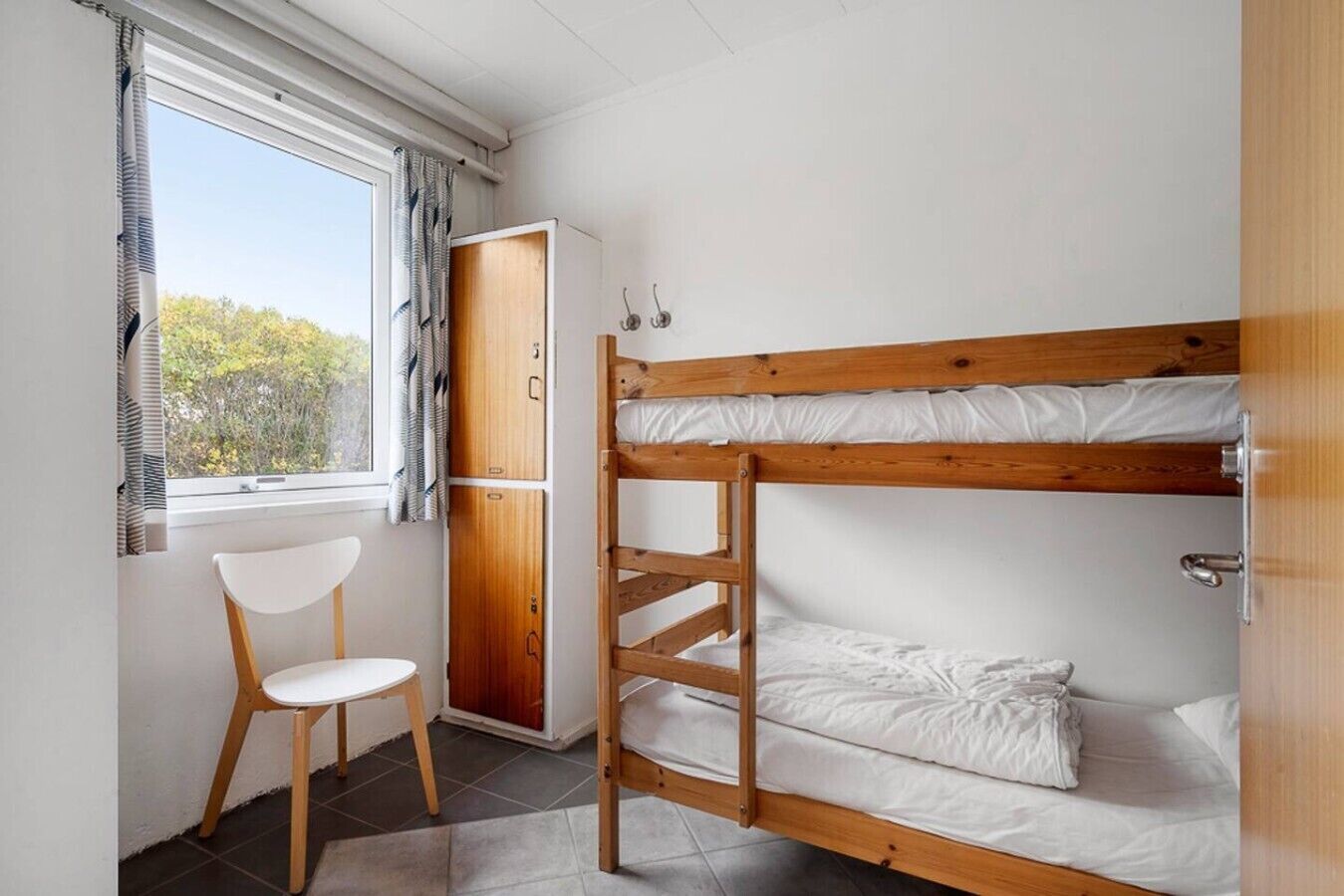 A bunk bed with white bedding, a wooden chair, a wardrobe, and a window with curtains.