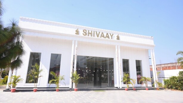 Un bâtiment blanc dont la façade porte le nom « SHIVAAY », avec de grandes fenêtres et des plantes en pot devant l’entrée.