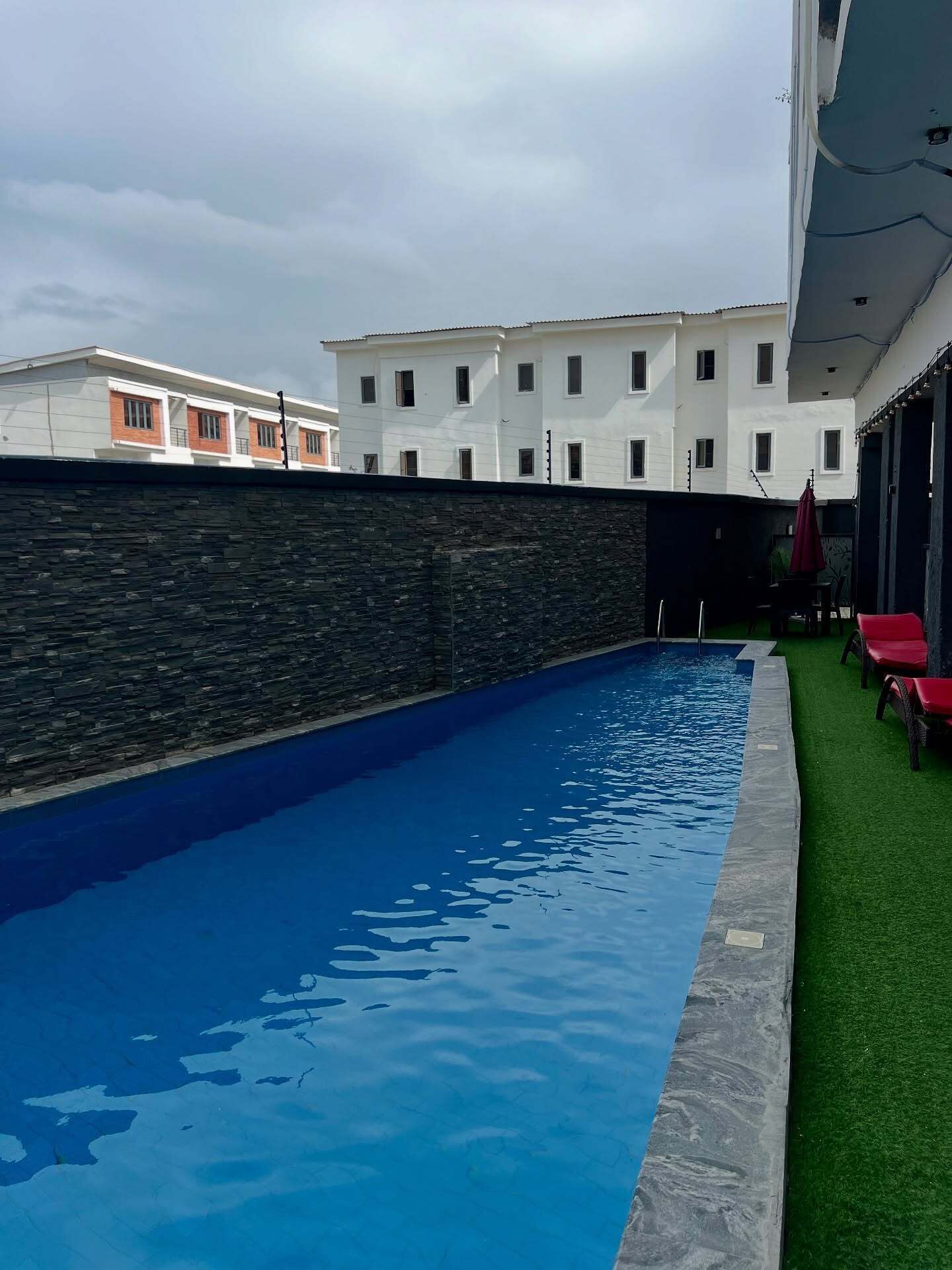 A swimming pool with a stone wall and a green lawn area with red chairs.
