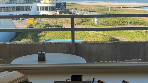 A dining area with a view of the beach, a sign for 'CUNDUNER', a coffee pot, a basket of bread, and a table with a book and mug. - Exclusive apartment with sea view in Haus Seeblick! - Lake view (Borkum)