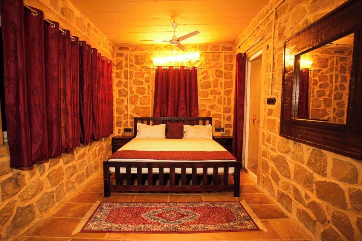 Un dormitorio con paredes de piedra, una cama de madera, cortinas rojas, un espejo y un ventilador de techo.