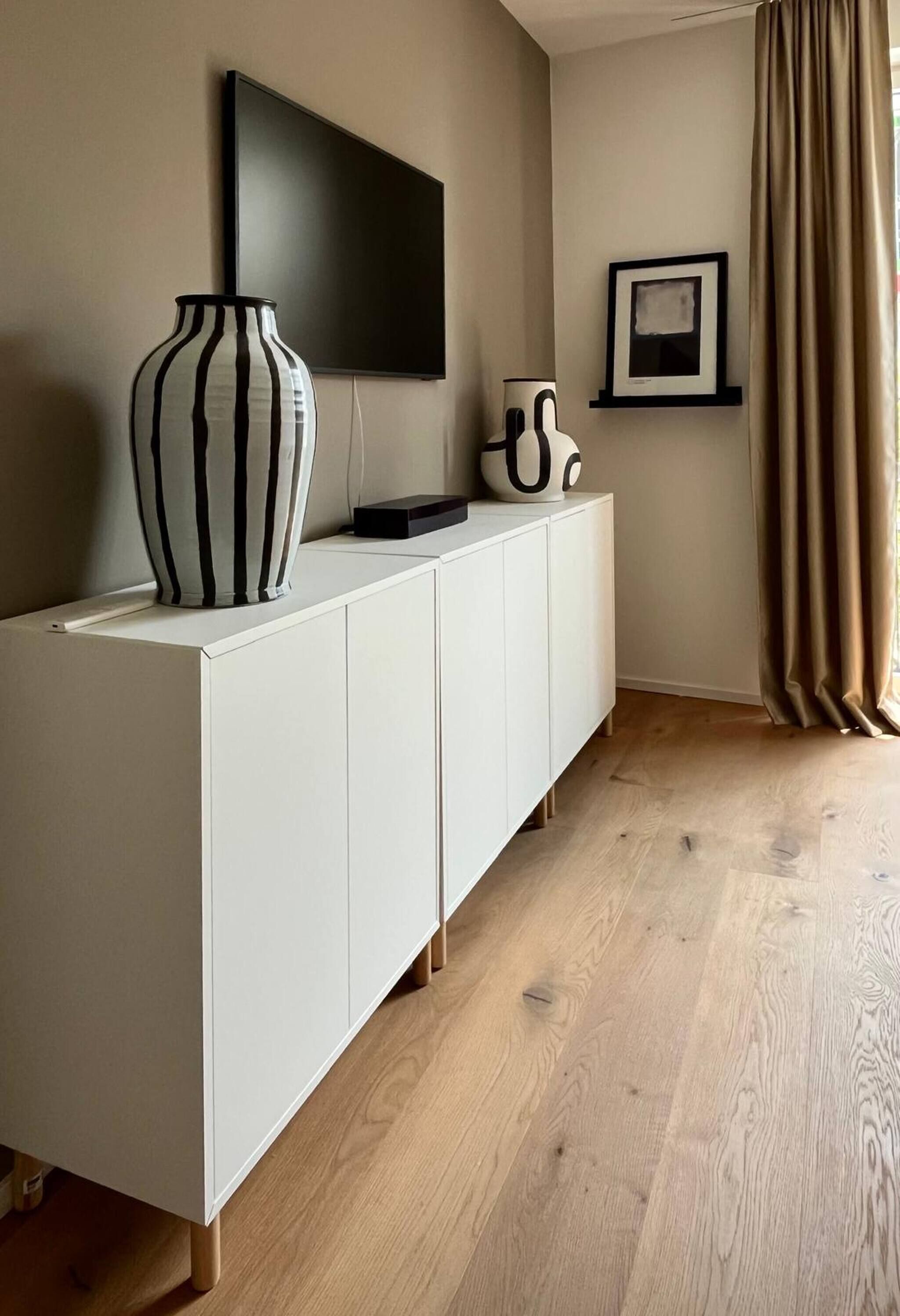 Een moderne woonkamer met een witte kast, een zwart-wit gestreepte vaas, een flatscreen televisie, een ingelijste foto en een bruin gordijn.