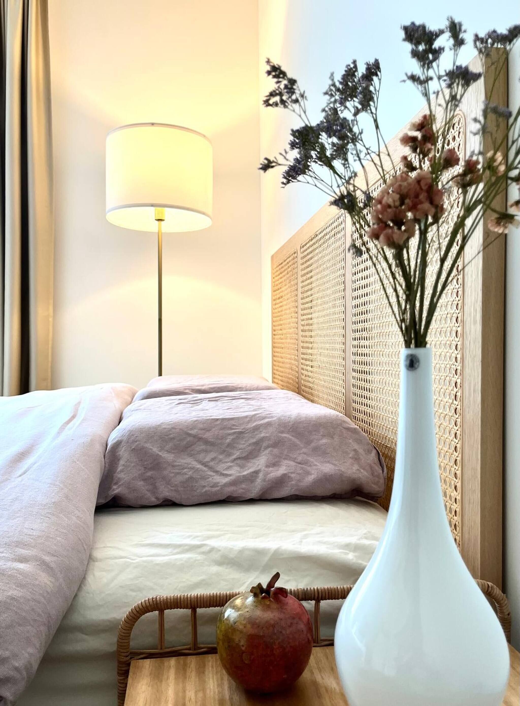 A hotel room with a bed, a lamp, a wicker basket, and a vase with flowers.