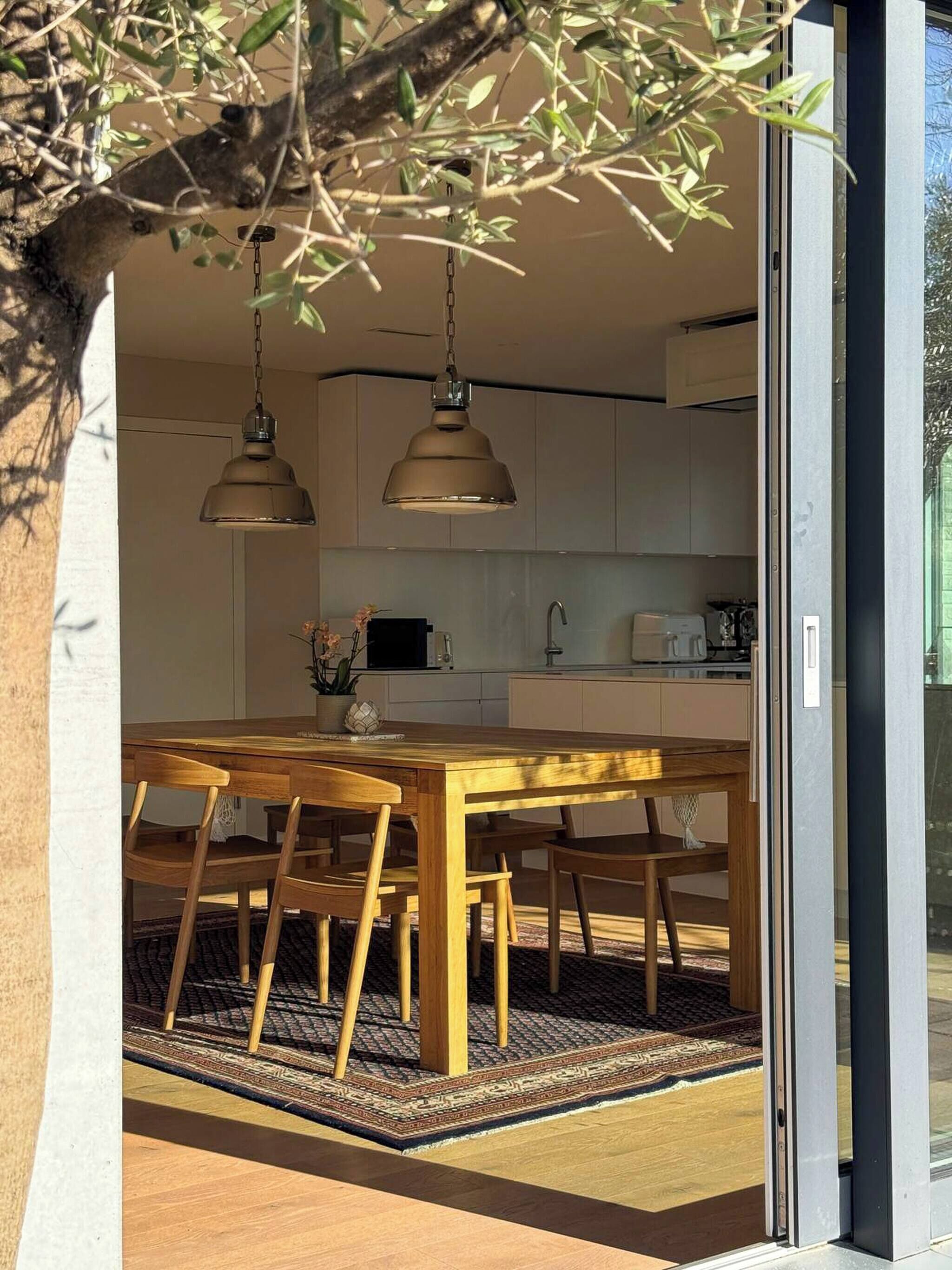 A modern kitchen with pendant lighting, a dining table with chairs, and a view through a glass door.
