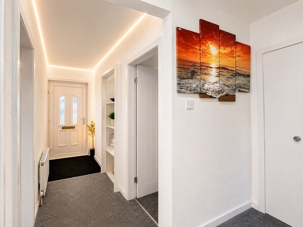 A hallway with a white door, a shelf with a potted plant, and a wall-mounted artwork depicting a sunset over the ocean.