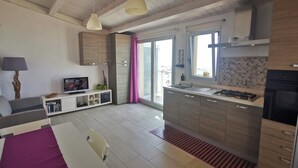 A modern kitchen with a central island, a dining area with a table and chairs, and a living area with a sofa and TV. - Appartamento Tramonto sul Mare (Caucana)