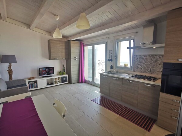 Eine moderne Küche mit einer zentralen Kochinsel, ein Essbereich mit Tisch und Stühlen sowie ein Wohnbereich mit Sofa und Fernseher.