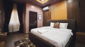 Quarto de hotel moderno com uma cama grande, uma mesa de cabeceira, ar condicionado fixado na parede e uma janela com cortinas leves.