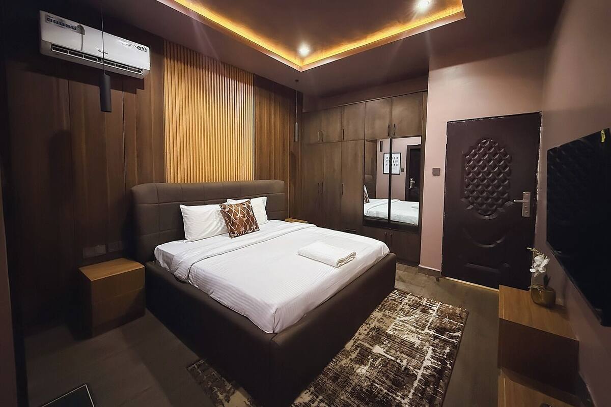 Una habitación de hotel moderna con una cama grande, paneles de madera, un armario empotrado y una unidad de aire acondicionado instalada en el techo.