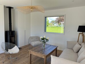 Ein modernes Wohnzimmer mit Kaminofen, einem Esstisch mit Blumenvase, einem grauen Sessel und einer Couch.