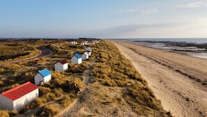 A coastal area with a row of small, white beach huts with colorful roofs, a sandy beach, and a road curving through the landscape. - New wooden house less than 5 minutes from the beach (Gouville-sur-Mer)