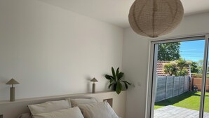 A bedroom with a large bed, a ceiling light fixture, and a view of a backyard through a sliding glass door. - New wooden house less than 5 minutes from the beach (Gouville-sur-Mer)