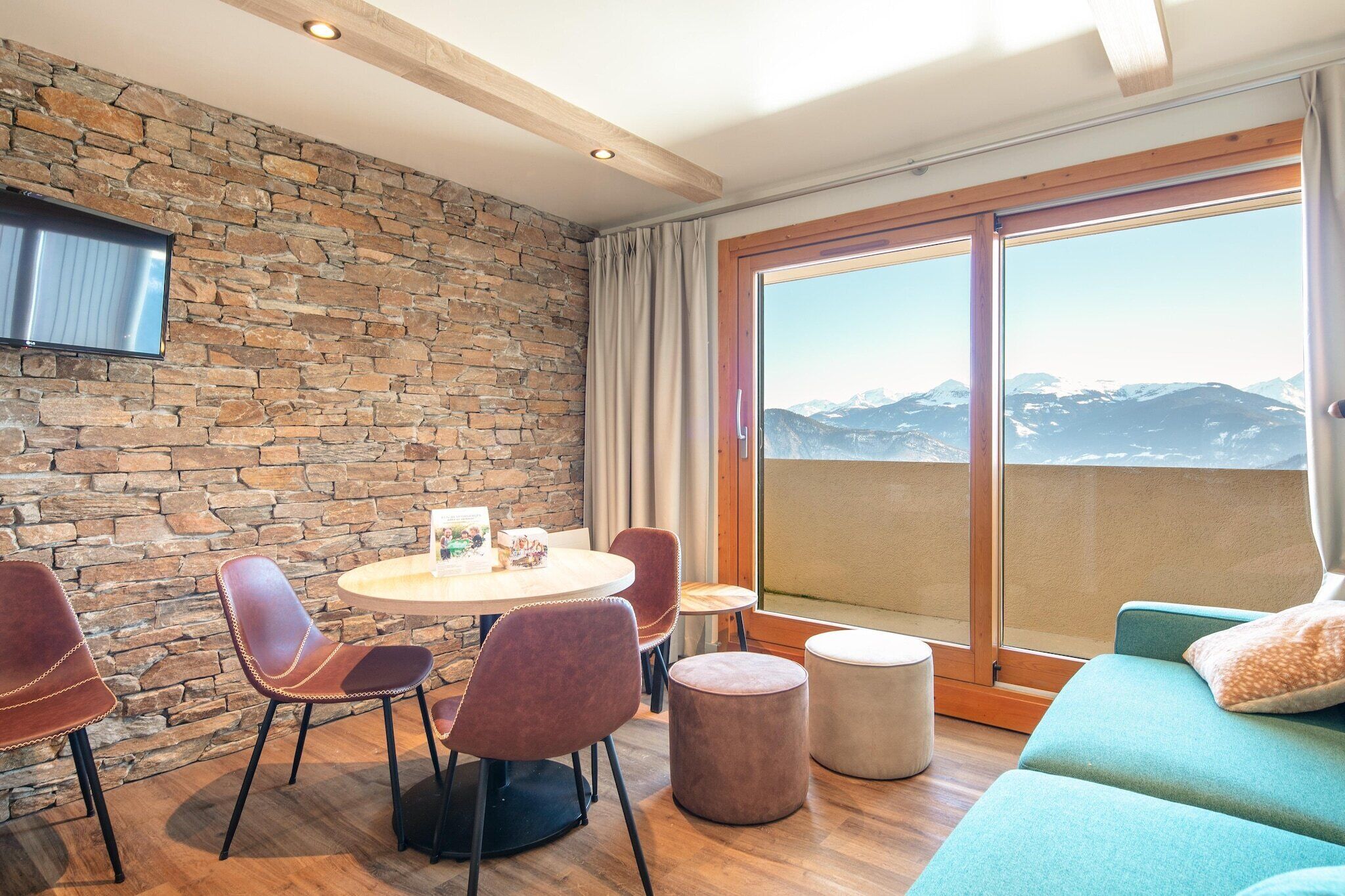 Une pièce avec un mur en pierre, une table ronde entourée de chaises, un canapé, un téléviseur fixé au mur et une grande fenêtre donnant sur la montagne.