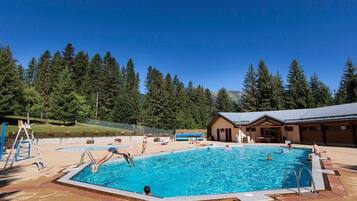 Une piscine avec une terrasse en bois, un petit bâtiment et, en arrière-plan, une colline boisée.