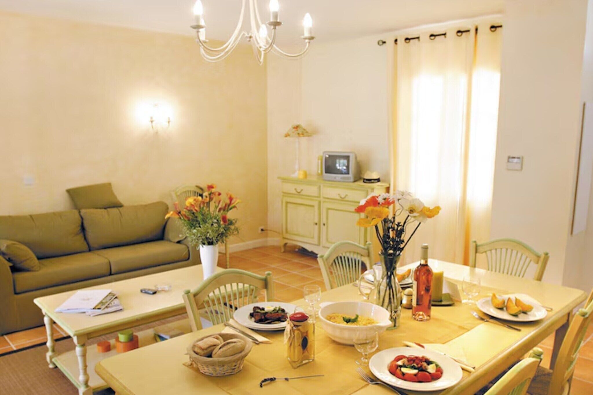 A dining area with a table set for two, a sofa, a TV, and a vase with flowers.