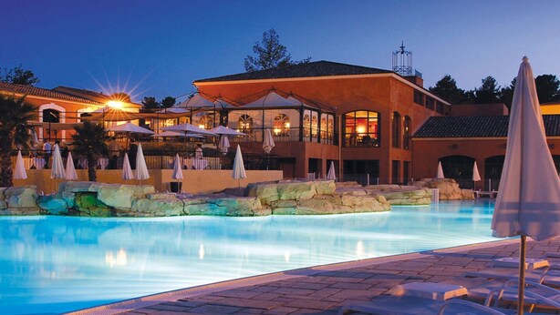 Ein Schwimmbecken mit Liegestühlen, Sonnenschirmen und einem mehrstöckigen Gebäude mit Außenbereich.