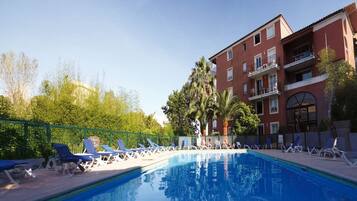 Piscina com espreguiçadeiras, um edifício com varandas e vegetação ao fundo.