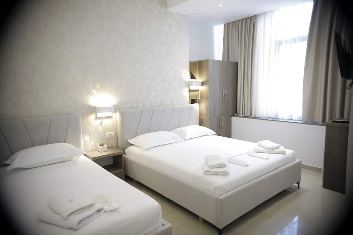 A hotel room with two beds, a wardrobe, a nightstand with a lamp, and a window with curtains.