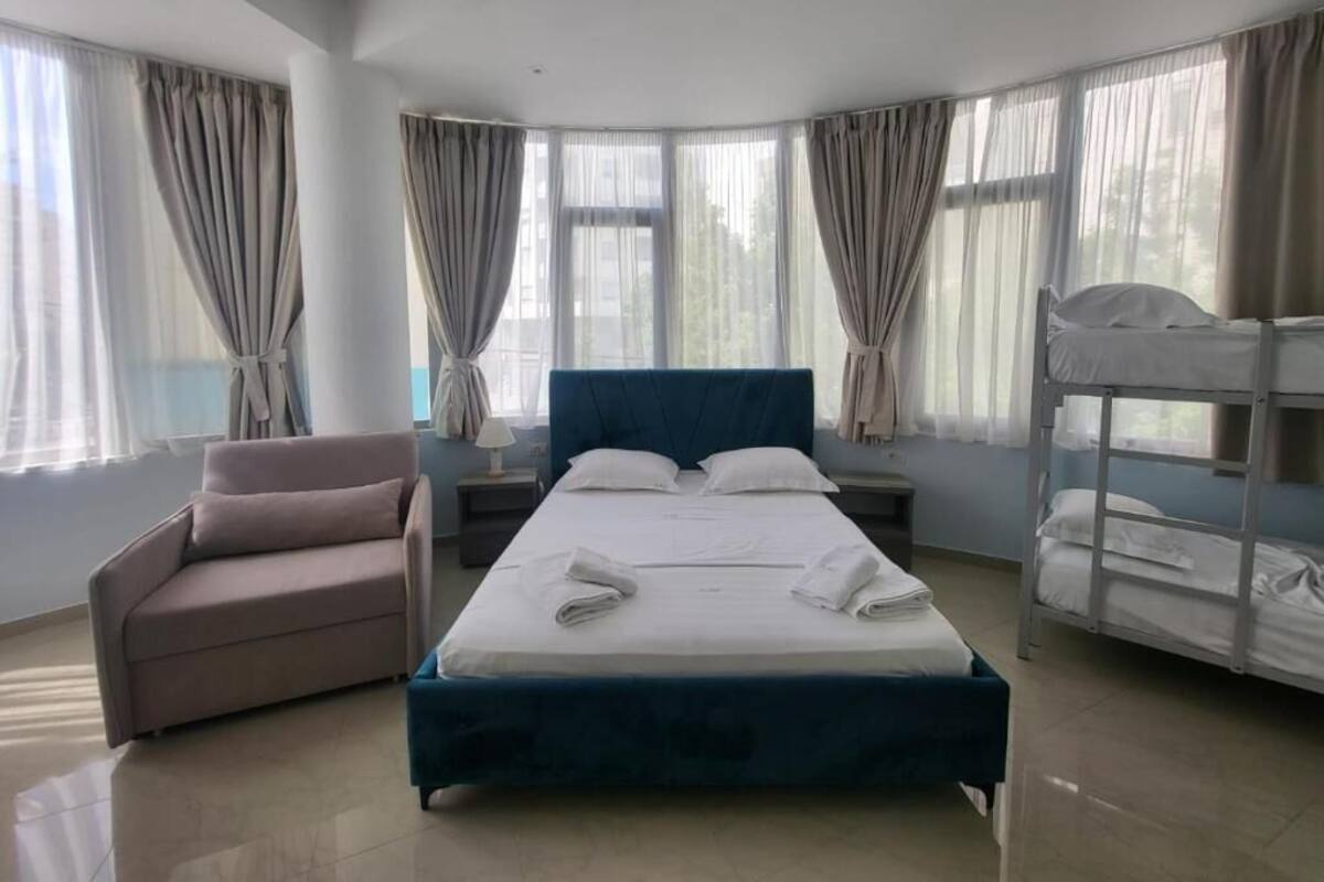 A hotel room with a bed, a chair, a bunk bed, and large windows with curtains.