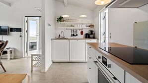 A modern kitchen with white cabinetry, a wooden countertop, and a built-in oven. There is a dining area with a table and chairs, and a TV mounted on the wall. - Sct. Knudsborg · Cottage 12H · Sea view (Middelfart)