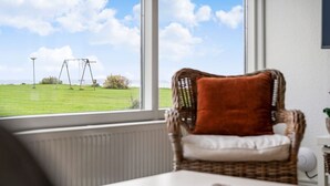 A wicker chair with a cushion, a white table, and a view of a grassy field with a swing set through a window. - Sct. Knudsborg · Cottage 12H · Sea view (Middelfart)