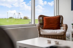 Ein Korbstuhl mit Kissen, ein weißer Tisch und Blick auf ein Wiesenfeld mit Schaukeln durch ein Fenster.