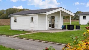A small, single-story mobile home with a covered porch, a small attached garage, and a grassy yard. - Sct. Knudsborg · Cottage 12H · Sea view (Middelfart)