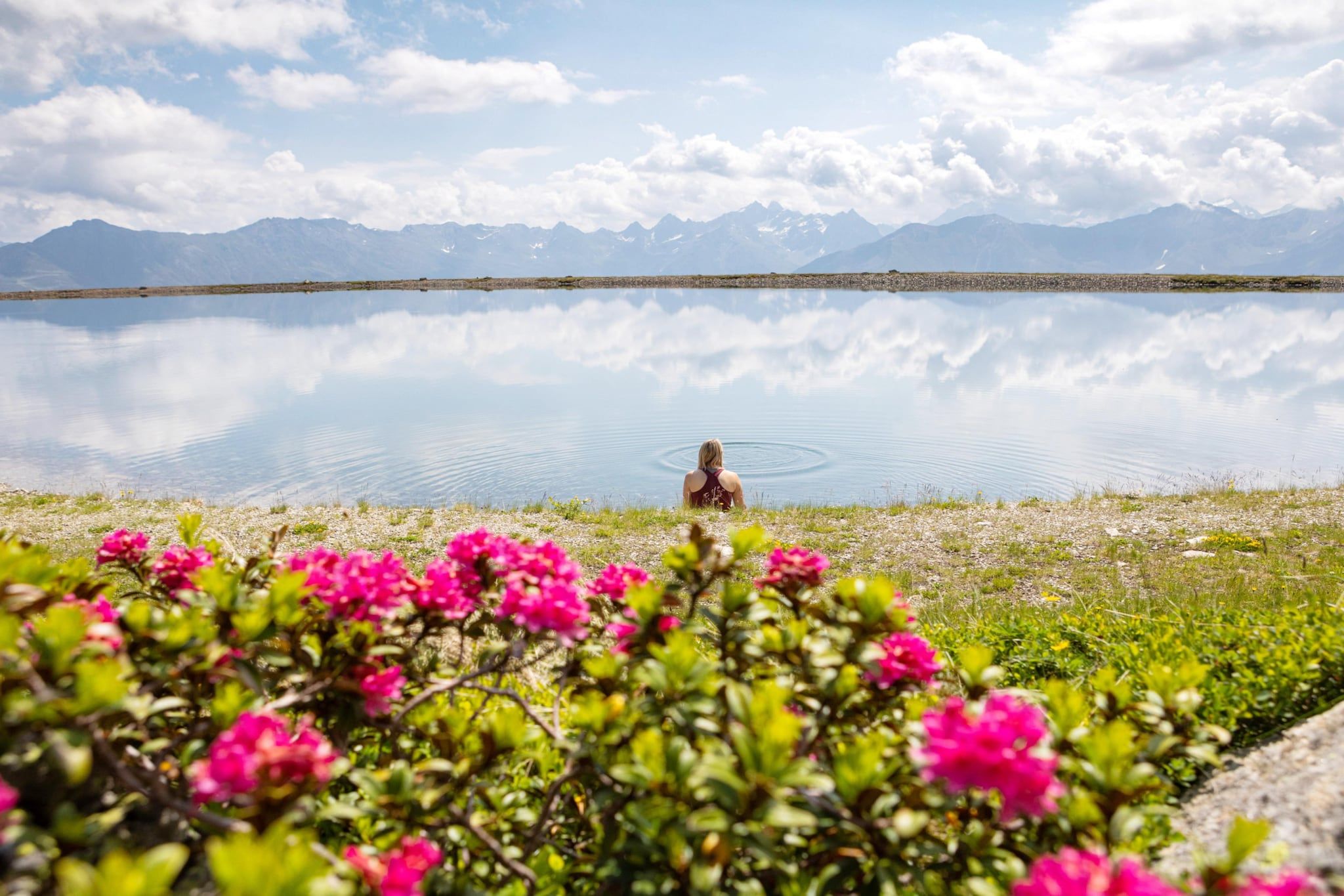 Eine Person sitzt an einem ruhigen See, im Hintergrund Berge.
