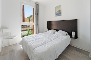 Ein Schlafzimmer mit Bett, Stuhl, Nachttisch, Lampe, einem Fenster mit Vorhängen und einem Wandbild.
