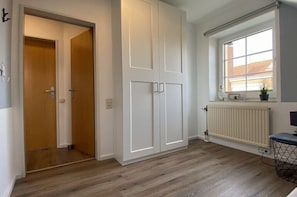 A room with a wooden floor, white walls, a radiator, a window with a view, and a door leading to another room. - Holiday apartment in Greetsiel with Wi-Fi (Greetsiel)