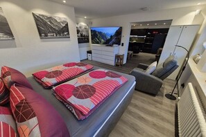 A modern living room with a sofa, a flat-screen TV, and a kitchen area in the background. - Holiday apartment with 1 bedroom (Oberstdorf)