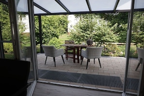 Ein verglaster Patio mit Tisch, Stühlen, einer Topfpflanze und Blick auf einen Garten.