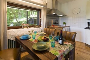 Ein Esstisch mit Tellern, Gläsern und einer Flasche, ein Fenster mit Blick auf Bäume und ein Küchenbereich mit Kochutensilien.