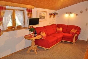 A cozy room with a red sofa, a small wooden table with a vase and snacks, a TV on the wall, and a window with curtains. - Holiday apartment with 1 bedroom (Wallgau)